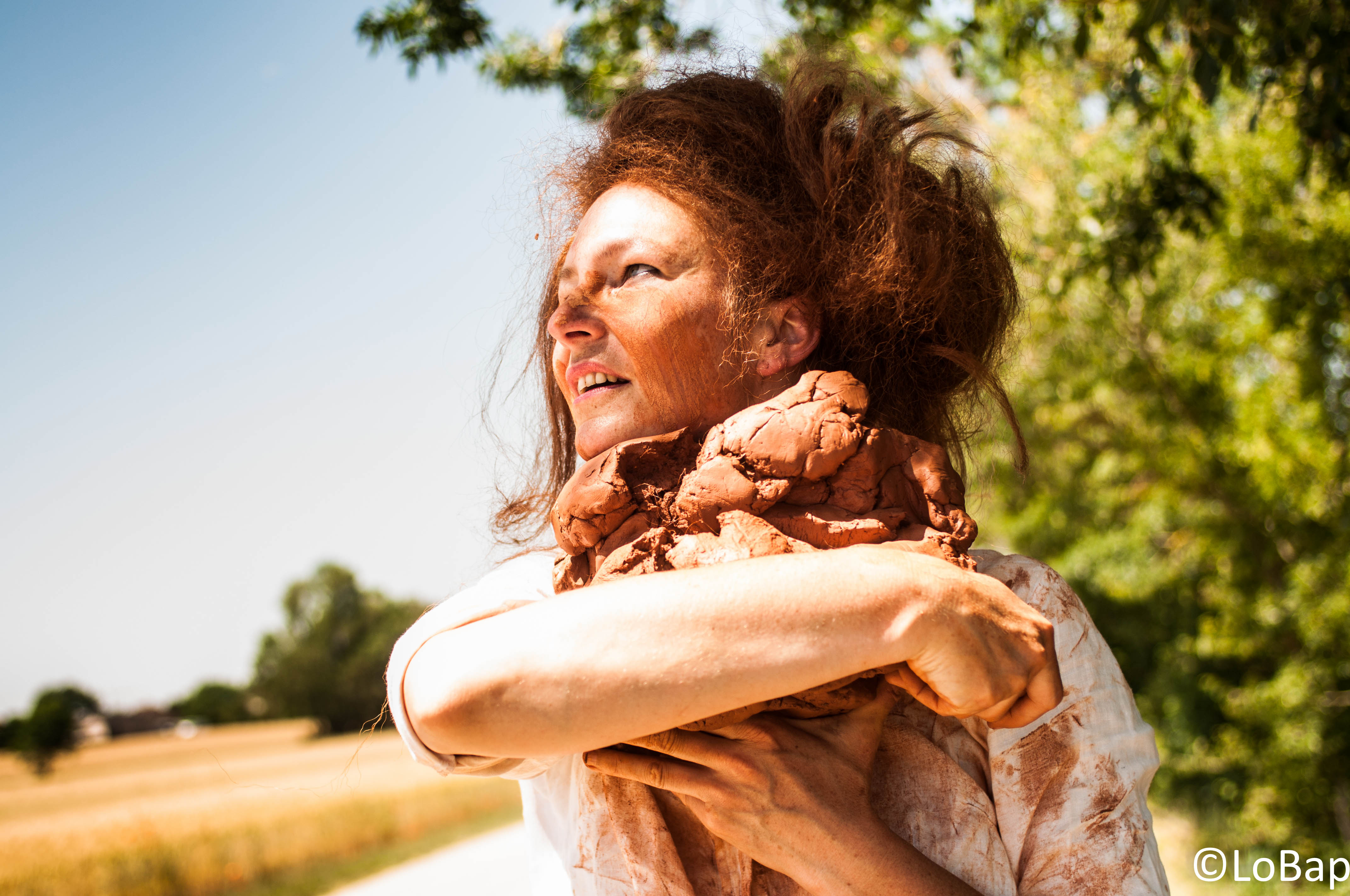 Femme avec des cheveux en désordre, souriant et tenant une sculpture en argile dans ses bras, sur un chemin ensoleillé entouré de verdure.